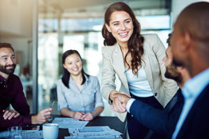 Young professional woman standing at a table of other professionals reaching to shake the hand of a man sitting at the table