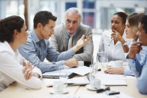 Business professionals engaged in a serious discussion around a conference table, with documents and laptops in front of them.